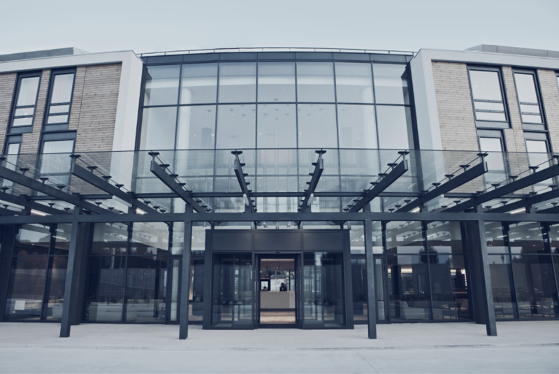 View of the steel and glass façade of the main entrance. The structure leaves room for the windows of the rooms with their wooden cladding.
