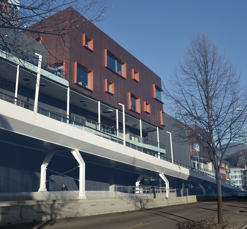 Nuovo blocco del centro commerciale, vista esterna. Le rampe portano i percorsi pedonali direttamente dentro il centro commerciale o al ponte che attraversa la via. I parapetti vetrati esterni usano lo stesso sistema di quelli interni con il corrimano in legno per dare continuità.
